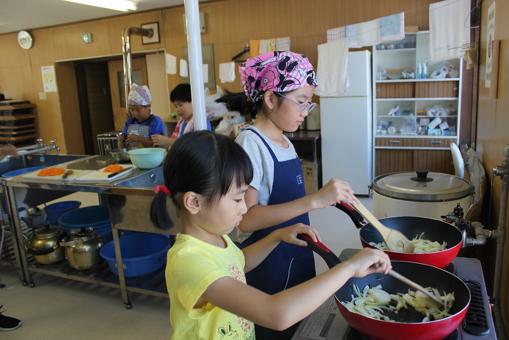 8月7日（火）生活改善センターにて、放課後児童クラブの一環で「炊事学習」が開催されました。毎年夏休み中に開催され、今年はみんなでピラフとパスタを作りました。上級生のお姉さんに教えてもらいながら料理していますね。